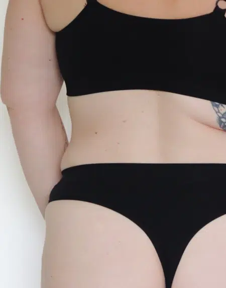 Personne portant une culotte noire grande taille sans couture, mettant en valeur la partie médiane des épaules jusqu'au haut des cuisses, avec un tatouage sur le bras gauche. Mode Grande Taille ajoute une touche inclusive à cette représentation élégante. Mode Grande taille