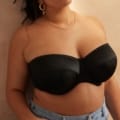 Une personne portant le Soutien gorge bandeau Dana noir et un jean bleu, parfait pour Mode Grande Taille, est représentée des épaules à la taille, debout près d'un mur beige. Mode Grande taille