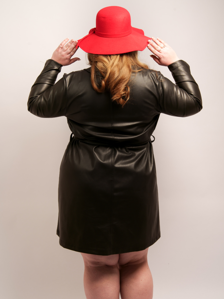 Une personne en tenue de cuir noir et chapeau rouge, vue de dos, ajuste son chapeau sur un fond uni, adoptant le style chic de Mode Grande Taille. Mode Grande taille