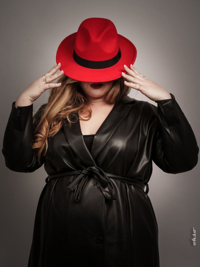 Personne portant un trench-coat en cuir noir tenant et couvrant partiellement son visage avec un chapeau rouge, posant sans effort sur un fond gris. Mode Grande Taille, sophistication à son meilleur. Mode Grande taille