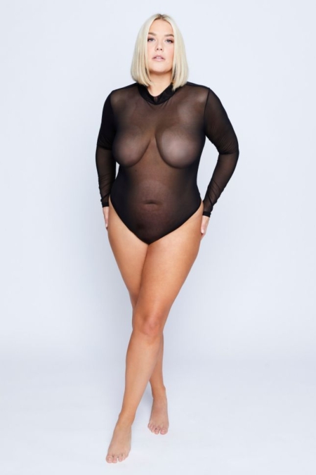 Une femme dans un body en mesh noir transparent à manches longues, incarnant la Mode Grande Taille, se tient avec confiance sur un fond uni gris clair. Mode Grande taille