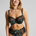 Une femme souriante porte le Soutien-gorge balconnet Esmé Ziggy, un ensemble de lingerie noir à motifs étoilés et détails en dentelle, posant sur un fond uni. Cet ensemble élégant de Mode Grande Taille met en valeur sa confiance et son style. Mode Grande taille