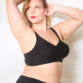 Une personne aux cheveux blonds porte un soutien-gorge à agrafes noir et un pantalon noir, posant avec un bras derrière la tête contre un mur blanc. Mode Grande taille