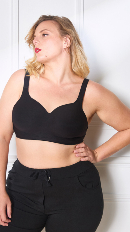 Personne portant un soutien-gorge à agrafes et un pantalon noir debout contre un mur blanc, regardant sur le côté avec une pose confiante. Mode Grande taille