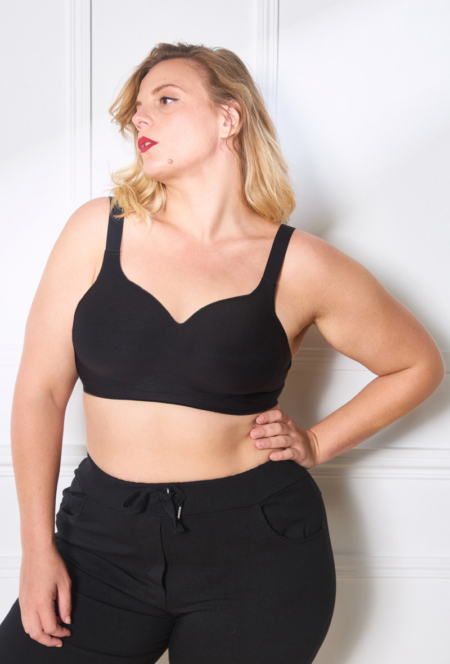 Personne portant un soutien-gorge à agrafes et un pantalon noir debout contre un mur blanc, regardant sur le côté avec une pose confiante. Mode Grande taille