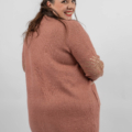 Femme aux cheveux longs souriant et regardant par-dessus son épaule, portant un pull rose et un pantalon foncé. Mode Grande taille