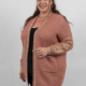 Femme souriante dans un cardigan rose avec des accents métalliques et un haut noir, posant sur un fond uni. Mode Grande taille