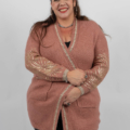 Une femme souriante aux cheveux longs, vêtue d'un cardigan rose avec des motifs dorés sur les manches, se tient debout sur un fond blanc. Mode Grande taille