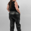 Femme en haut sans manches noir et pantalon en cuir noir, souriant et posant avec un bras levé, devant un fond gris. Mode Grande taille