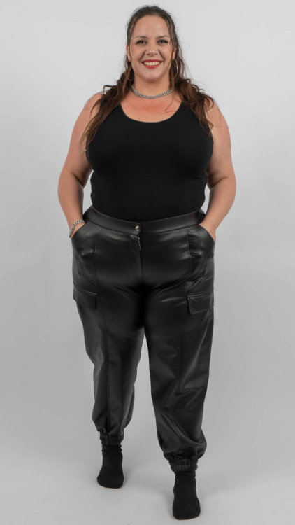 Une femme portant un débardeur noir et un pantalon en cuir noir se tient debout et sourit à la caméra, les mains dans les poches. Mode Grande taille