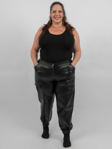 Une femme portant un débardeur noir et un pantalon en cuir noir se tient debout et sourit à la caméra, les mains dans les poches. Mode Grande taille