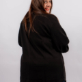 Femme portant un pull noir et un jean foncé, souriant par-dessus son épaule sur un fond blanc uni. Mode Grande taille