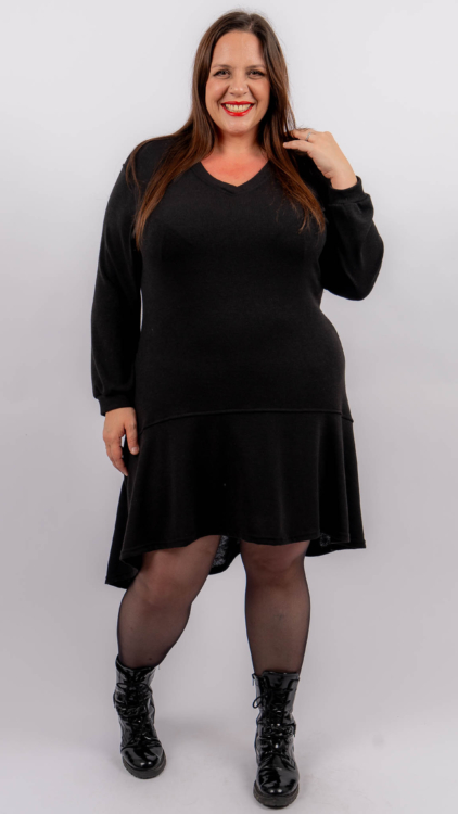 Femme souriante dans une robe noire à manches longues et des bottes noires, posant sur un fond blanc. Mode Grande taille