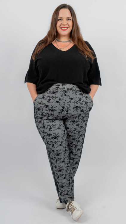 Une femme portant un haut noir et un pantalon à motifs, souriant avec ses mains dans ses poches, se tient debout sur un fond uni. Mode Grande taille