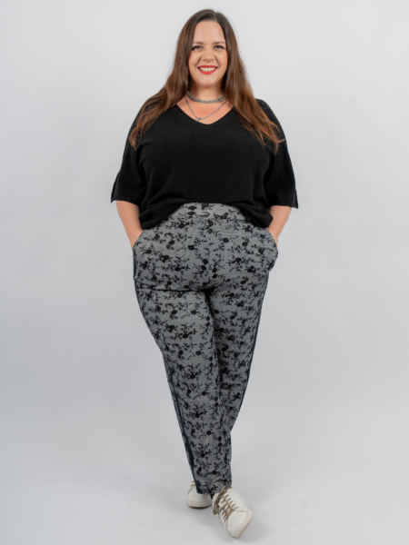 Une femme portant un haut noir et un pantalon à motifs, souriant avec ses mains dans ses poches, se tient debout sur un fond uni. Mode Grande taille