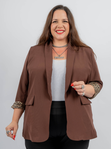 Une femme portant un blazer marron et un haut blanc sourit, un stylo à la main. Elle a les cheveux longs et un vernis à ongles rouge vif. Mode Grande taille