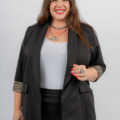 Une femme souriante aux cheveux longs portant un blazer noir, un haut blanc et des bijoux se tient debout sur un fond uni. Mode Grande taille