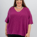 Une femme portant un haut violet et un pantalon noir sourit à la caméra, portant un collier et des bagues, les mains posées à ses côtés. Mode Grande taille