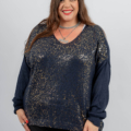 Femme souriante, portant un pull à paillettes bleu foncé et un pantalon noir, avec des ongles rouges et de longs cheveux bruns sur un fond uni. Mode Grande taille