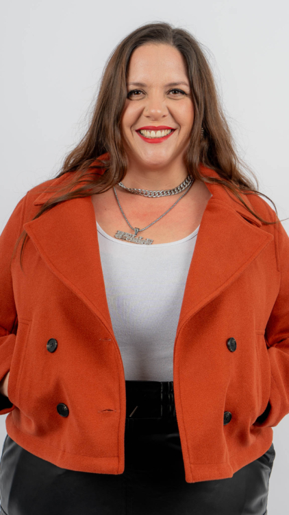 Une femme portant une veste rouge et un haut blanc sourit, les mains dans les poches, sur un fond uni. Mode Grande taille