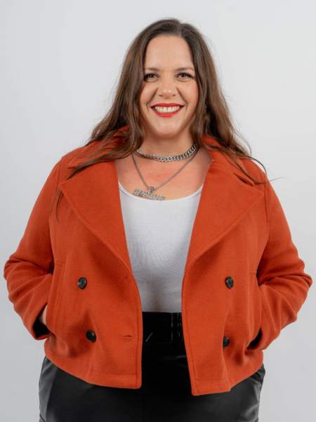 Une femme portant une veste rouge et un haut blanc sourit, les mains dans les poches, sur un fond uni. Mode Grande taille