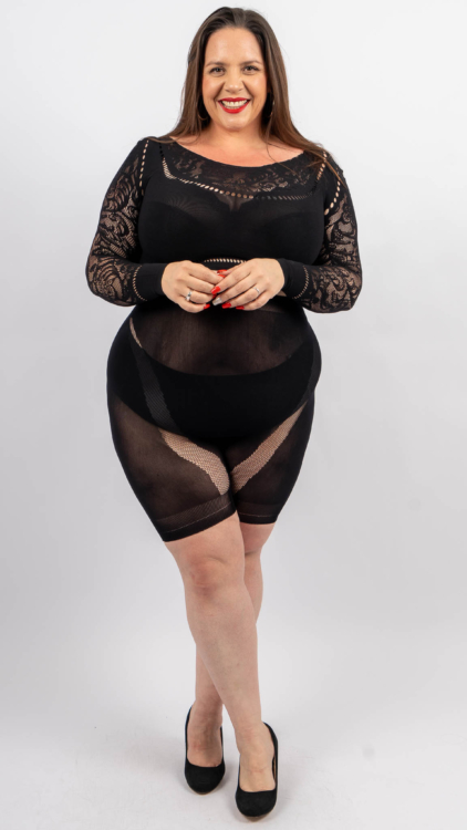 Une femme portant un haut noir en dentelle et un short noir assorti, debout et souriant, les mains jointes. Mode Grande taille