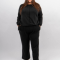 Une femme en tailleur-pantalon noir se tient debout avec confiance, les mains dans les poches, souriant à la caméra sur un fond uni. Mode Grande taille