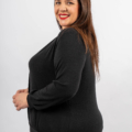 Femme souriante aux longs cheveux bruns portant un pull noir sur fond blanc, posant de profil latéral. Mode Grande taille