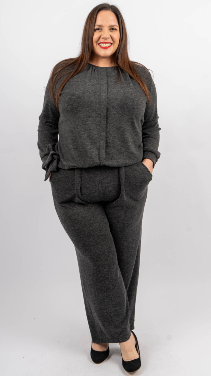 Femme portant un haut et un pantalon gris assortis, souriante, les mains dans les poches, debout sur un fond uni. Mode Grande taille