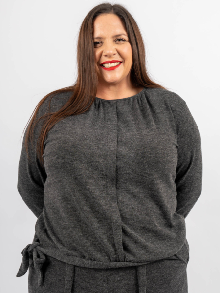 Une femme aux longs cheveux bruns, portant un haut gris foncé, sourit avec du rouge à lèvres rouge sur un fond blanc. Mode Grande taille