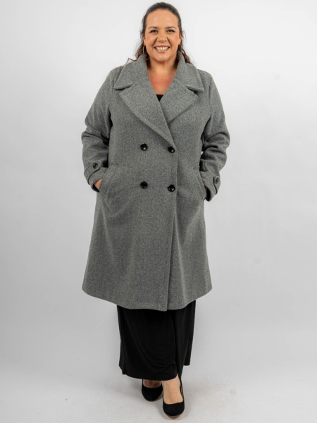 Femme souriante dans un long manteau gris avec les mains dans les poches, portant un pantalon noir et des talons. Mode Grande taille