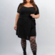 Femme en haut noir et jupe avec collants à motifs et bottes, souriant sur un fond uni. Mode Grande taille