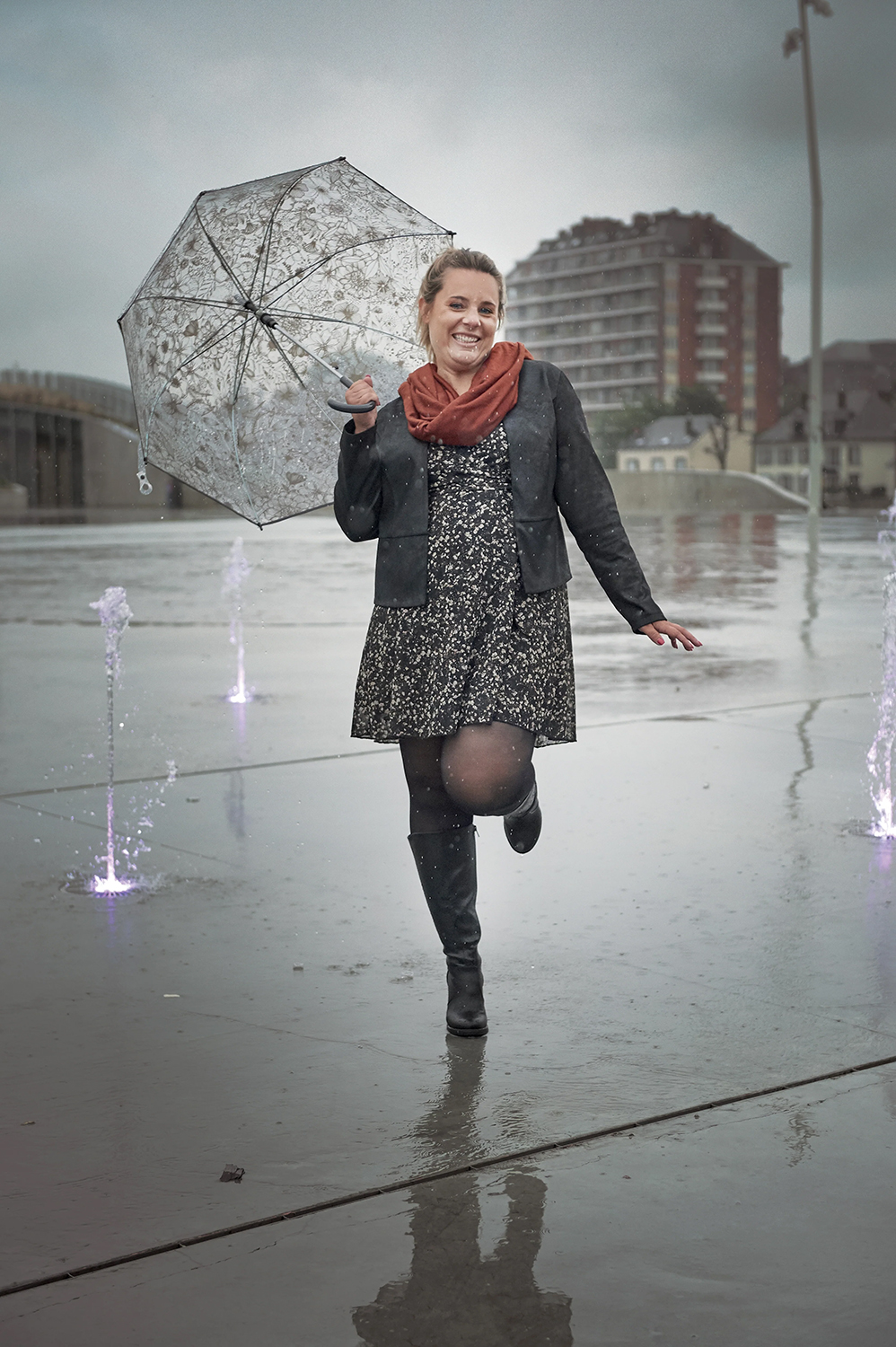 Femme souriante sous la pluie, tenant un parapluie transparent, portant une robe et un foulard, debout sur un trottoir mouillé avec des fontaines. Mode Grande taille