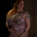 Une femme aux longs cheveux roux pose avec assurance, portant un haut Body Sequins Mauve scintillant et mettant en valeur ses tatouages. Mode Grande taille