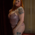 Une femme aux cheveux roux dans une tenue Body Sequins Mauve se tient debout avec confiance, mettant en valeur des tatouages sur son bras et sa cuisse. Mode Grande taille