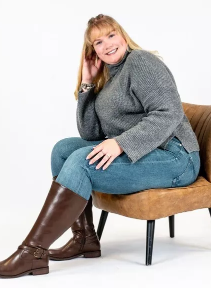 Personne souriante dans un chandail gris et un jean, assise sur une chaise marron, portant les bottes hautes à mollet large Lise 3XL Gabylou. Mode Grande taille