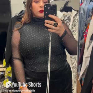 Une personne portant le « Haut à strass noir en mesh » prend un selfie devant un miroir dans une pièce ornée de vêtements et de décoration. Mode Grande taille