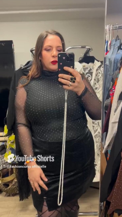 Une personne portant le « Haut à strass noir en mesh » prend un selfie devant un miroir dans une pièce ornée de vêtements et de décoration. Mode Grande taille