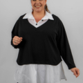 Femme souriante portant un pull noir sur une chemise blanche, debout sur un fond gris uni. Mode Grande taille