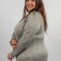 Une femme souriante dans un pull gris pose de côté sur un fond uni. Mode Grande taille