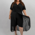 Femme en tenue noire avec superposition transparente, souriante et tenant le tissu, debout sur un fond uni. Mode Grande taille