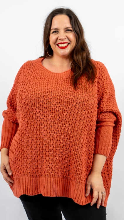 Une personne portant un pull orange et un pantalon noir sourit sur un fond blanc uni. Mode Grande taille