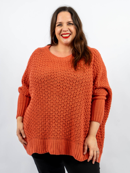 Une personne portant un pull orange et un pantalon noir sourit sur un fond blanc uni. Mode Grande taille