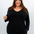 Femme souriante dans un pull noir aux cheveux longs, posant sur un fond uni. Mode Grande taille