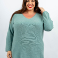 Femme portant un pull vert souriant et touchant ses cheveux d'une main sur un fond blanc uni. Mode Grande taille