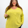 Femme souriante, portant un pull jaune vif et un pantalon noir sur un fond blanc uni. Mode Grande taille