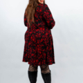 Femme portant une robe fleurie rouge et des bottes noires, souriant tout en regardant par-dessus son épaule sur un fond blanc. Mode Grande taille