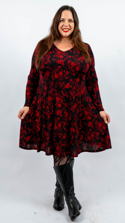 Femme en robe noire à motif floral rouge, souriante, debout sur un fond uni, portant des bottes noires. Mode Grande taille