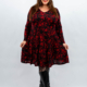 Femme en robe noire à motif floral rouge, souriante, debout sur un fond uni, portant des bottes noires. Mode Grande taille