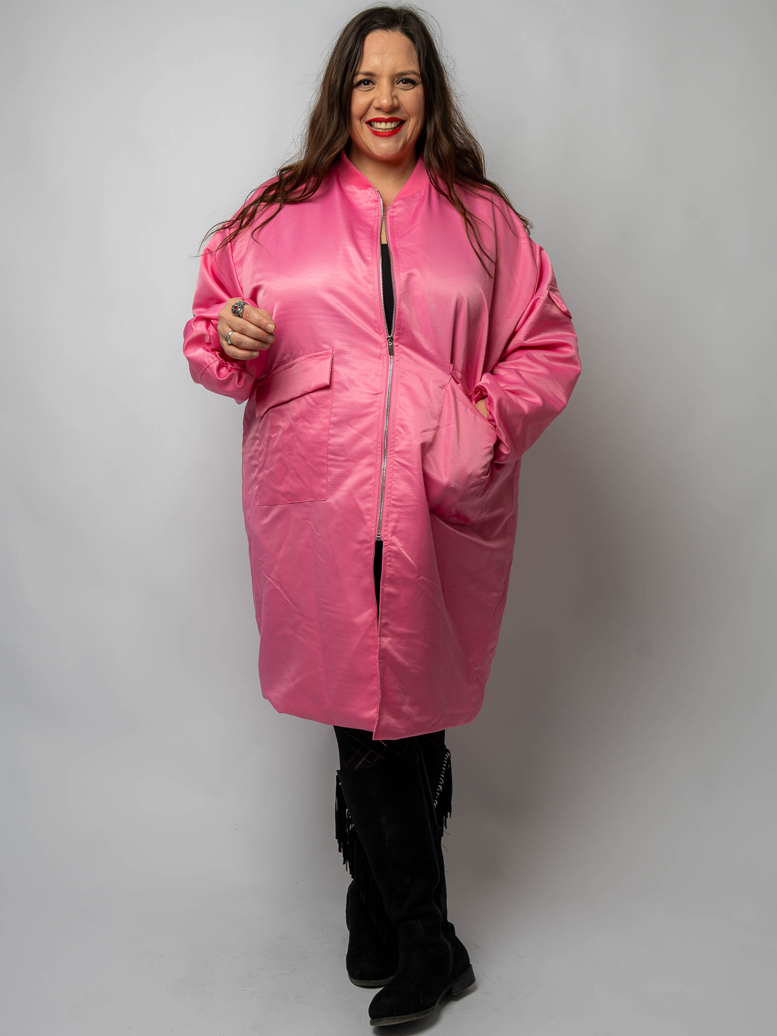 Une femme vêtue d'un long manteau rose, souriante, une main dans une poche, se tient debout sur un fond uni. Mode Grande taille